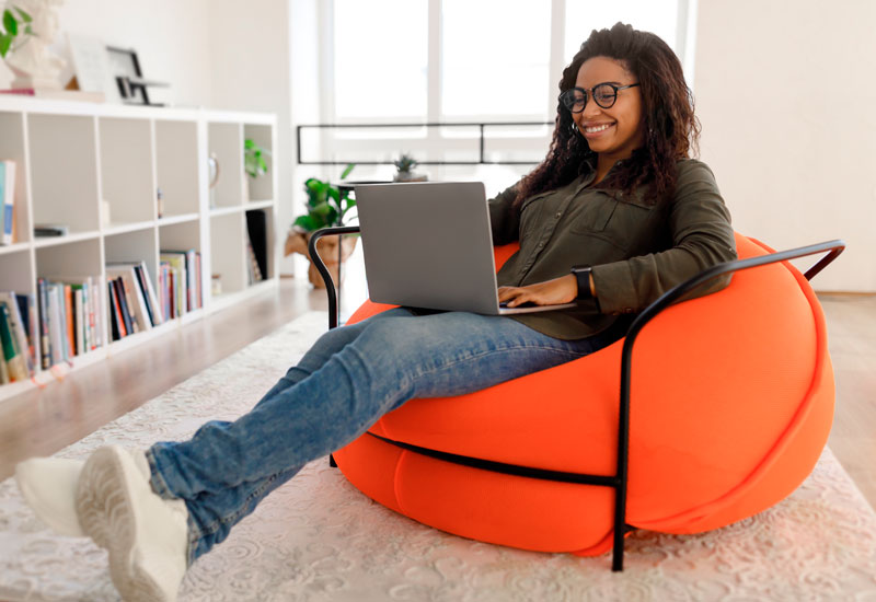 Empreendedora negra sentada num puff macio sorri enquanto utiliza seu notebook em um ambiente claro e sóbrio.